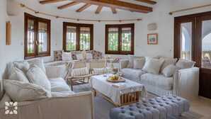 A spacious living room with a high ceiling, wooden beams, and large windows. There are multiple seating areas with white furniture and a central coffee table. - Villa PERLA by Abahana Villas (Teulada)