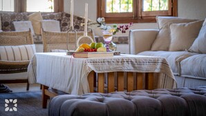 A cozy living room with a wooden table set with fruits and candles, wicker furniture, and a view of greenery through the window. - Villa PERLA by Abahana Villas (Teulada)
