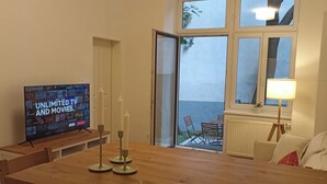 Una sala de estar con un televisor mostrando un servicio de streaming, una mesa de comedor de madera y vistas al exterior a través de un ventanal.