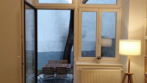 Una puerta abierta que da a un área de estar exterior con una mesa y sillas, un radiador y una lámpara de pie en una habitación con suelo de madera.