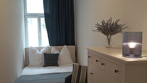 Un dormitorio con cama, una silla, un aparador, un florero con plantas secas y una lámpara.