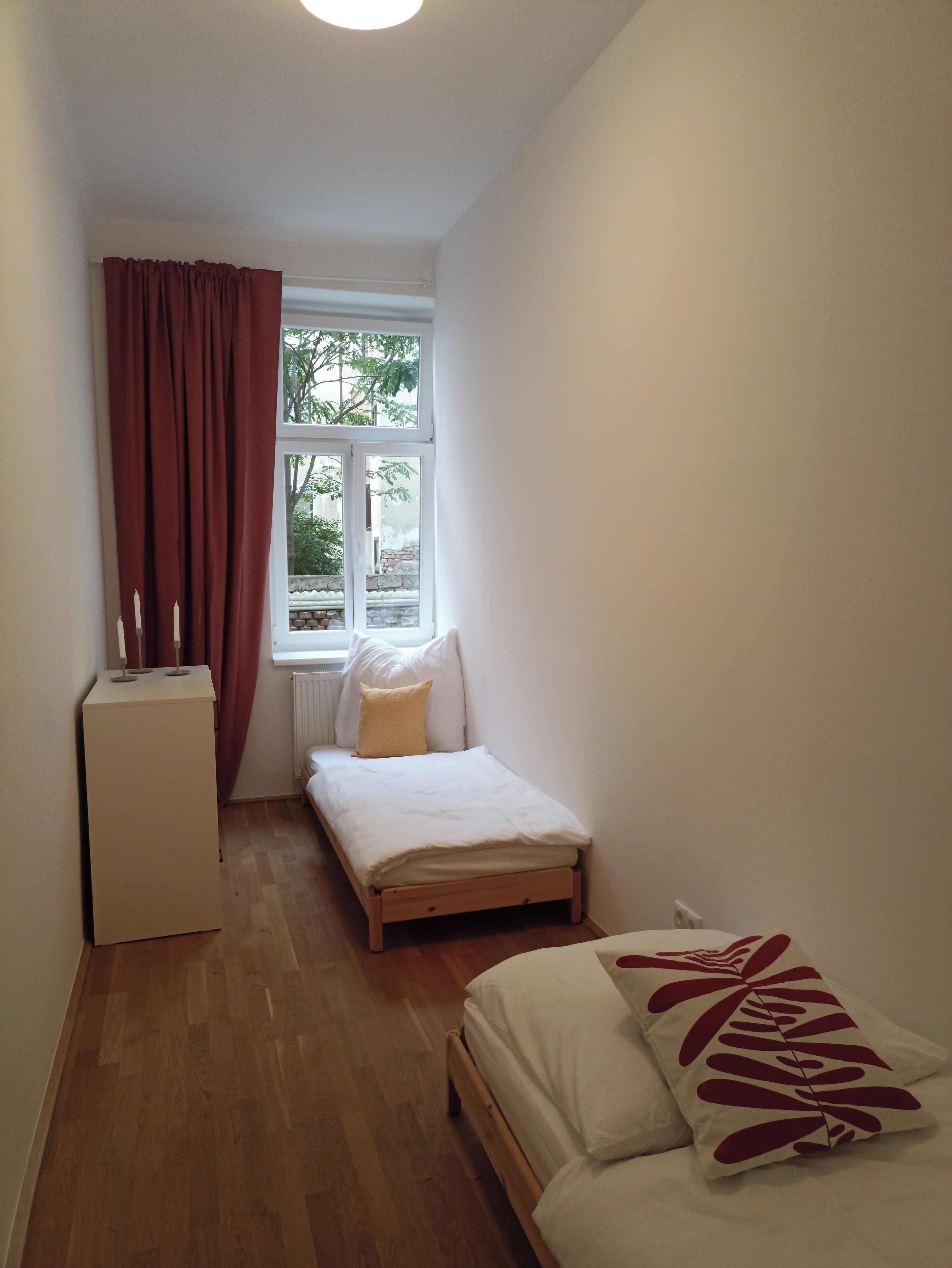 A small, well-lit room with a single bed, a small desk, and a window with curtains.