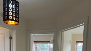 Un couloir mène à une chambre à coucher qui contient un lit, une table de chevet avec une lampe, et offre une vue sur l’extérieur par une fenêtre.