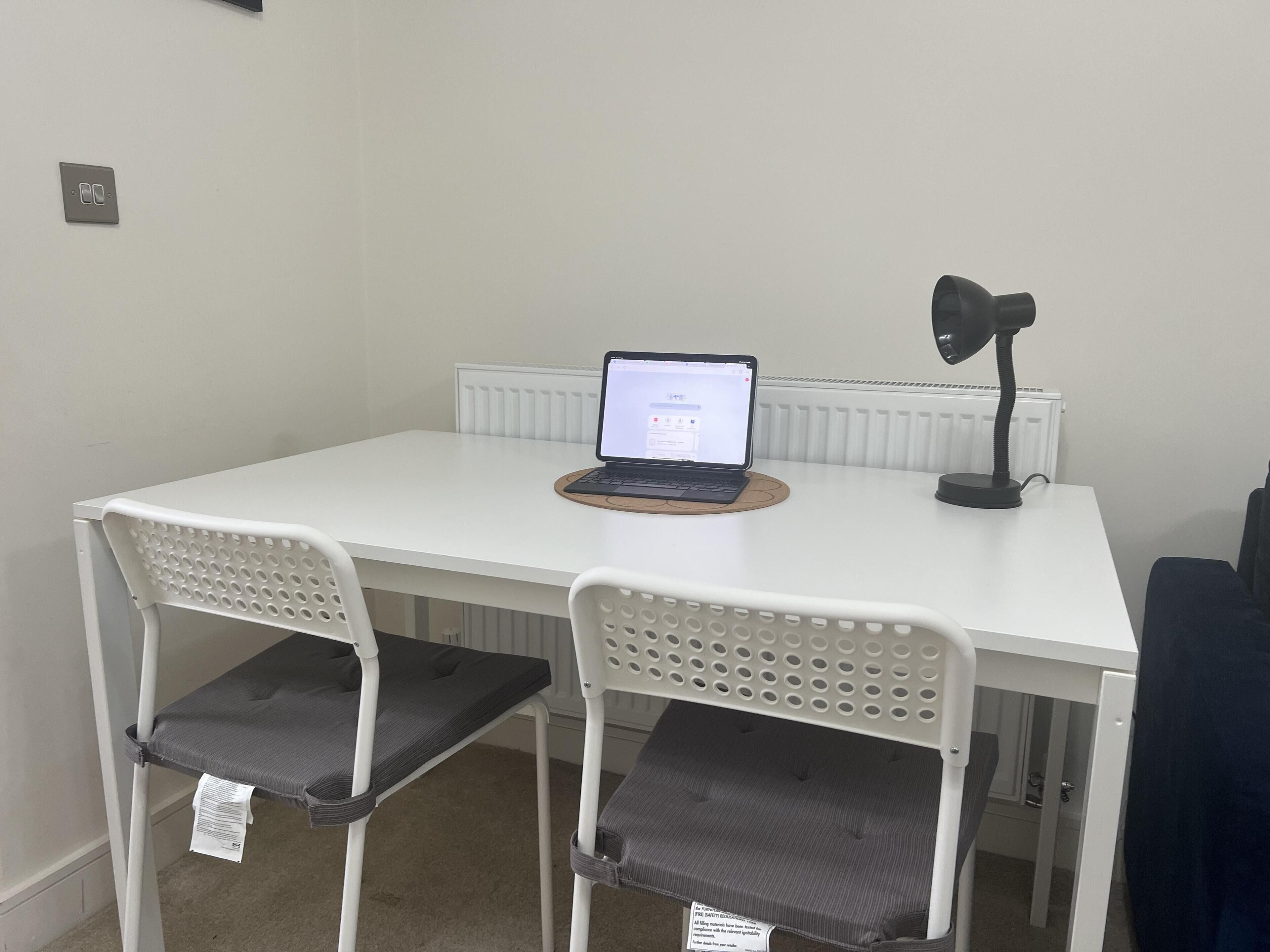 Una mesa pequeña y blanca con una computadora portátil y una lámpara de escritorio. Hay dos sillas blancas con respaldos perforados.