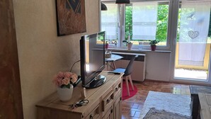 Een kamer met een houten bureau, een computer, een vaas met bloemen, een groot raam met gordijnen en een kamerbreed tapijt.