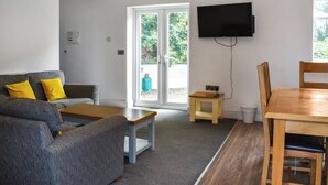 A living room with a sofa, coffee table, and a dining table with chairs. There is a television mounted on the wall and a window with a view of greenery outside. - 3 bedroom accommodation in Llanilar, near Aberystwyth (Llanilar, near Aberystwyth)