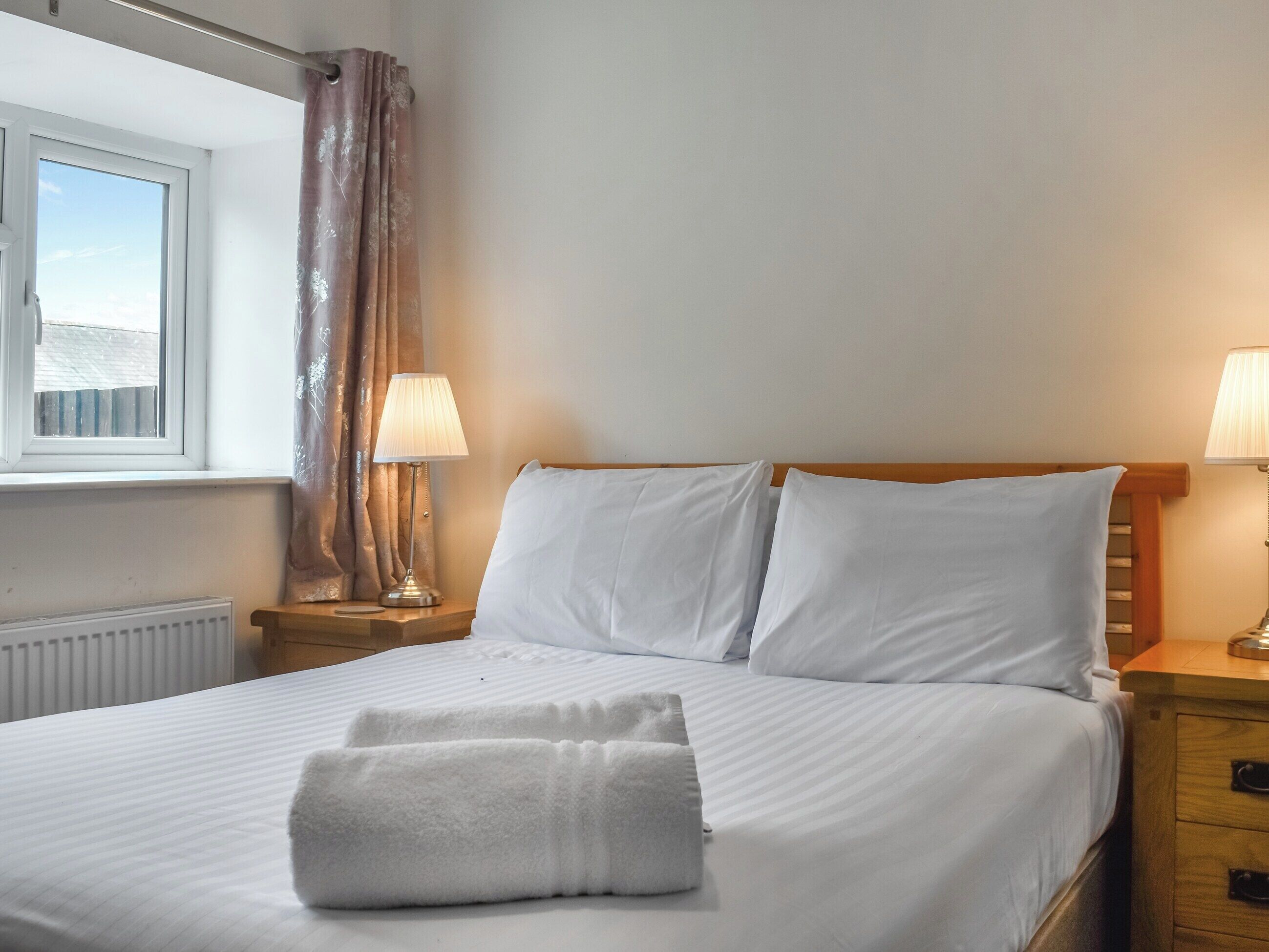 A neatly made bed with white linens and two pillows, a window with a view of a building, and a wooden nightstand with a lamp.
