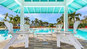 A covered pool area with white chairs and a table, surrounded by palm trees and a clear blue sky. - New 2 Bedroom + Den, 2 Bath Home w/2 Car Garage 10 Min from Downtown Naples (Naples)