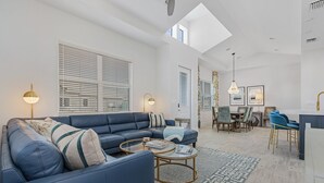 A modern living room with a blue sofa, a glass coffee table, and a dining area with blue chairs. - New 2 Bedroom + Den, 2 Bath Home w/2 Car Garage 10 Min from Downtown Naples (Naples)