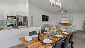 A modern kitchen with stainless steel appliances, a dining area with a wooden table set for four, and a living area with a sofa and a TV.