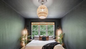 Et soveværelse med et stort vindue, en seng med sort sengegavl og en hængende lampe.