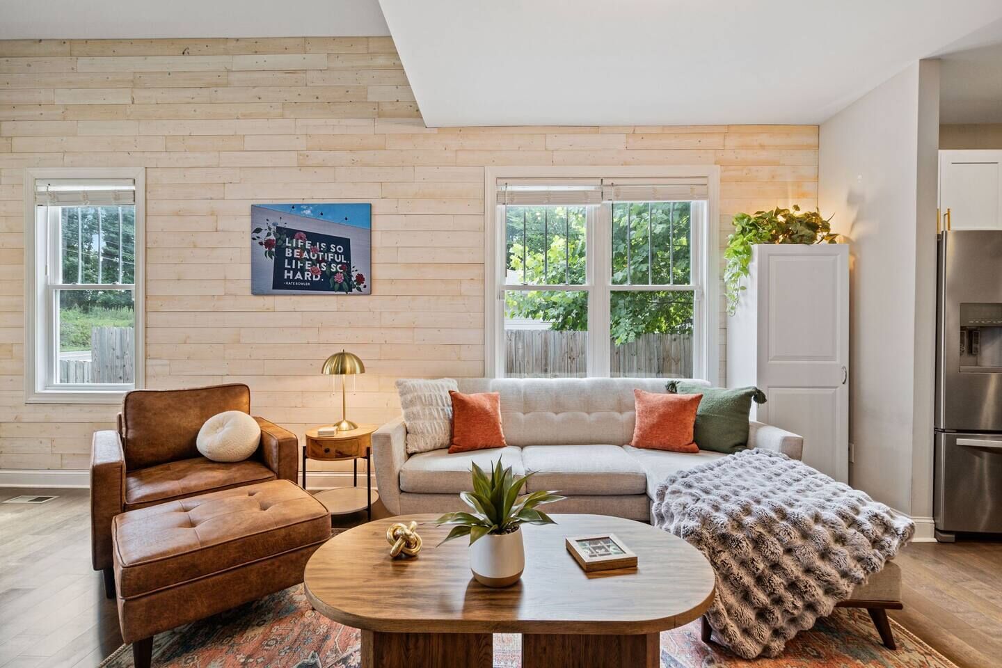 Een woonkamer met een bruine leren bank, een witte bank met kleurrijke kussens, een ronde houten salontafel met een plant en een boek, en een groot raam met uitzicht op groen.