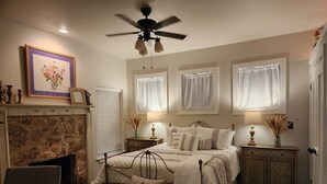 Une chambre à coucher avec une cheminée, un lit agrémenté de coussins décoratifs, un ventilateur de plafond et une table de chevet avec une lampe.