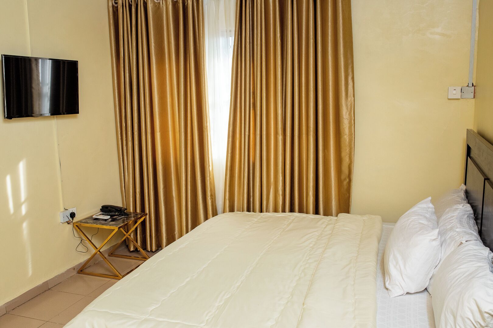 A hotel room with a bed, a TV mounted on the wall, and a desk with a phone and a lamp.
