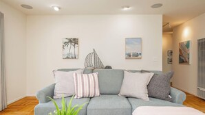 A living room with a grey sofa, a coffee table, and a potted plant. There are wall-mounted pictures and a window with blinds. - Entire Oceanview Coastal Retreat in Pacifica (Pacifica)