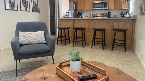 A living room with a grey armchair, a brown ottoman, a tray with a plant and remote control, and a kitchen with wooden cabinets and a microwave. - Charming one bedroom in a gated community near Sabino Canyon with heated pool. (Tucson)
