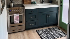Une cuisine avec des meubles blancs, un four en acier inoxydable, une planche à découper en bois et un tapis à motifs.