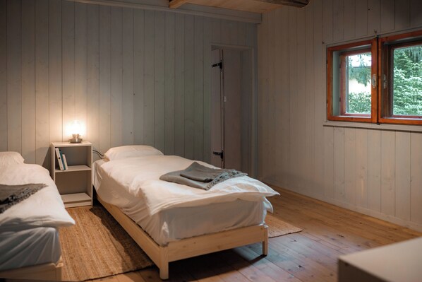 Un dormitorio con dos camas individuales, una mesita de noche con lámpara, una puerta y una ventana con vistas a árboles.