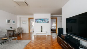 A modern living room with a large flat-screen TV, a dining area with a white table and chairs, and a hallway leading to a bedroom.