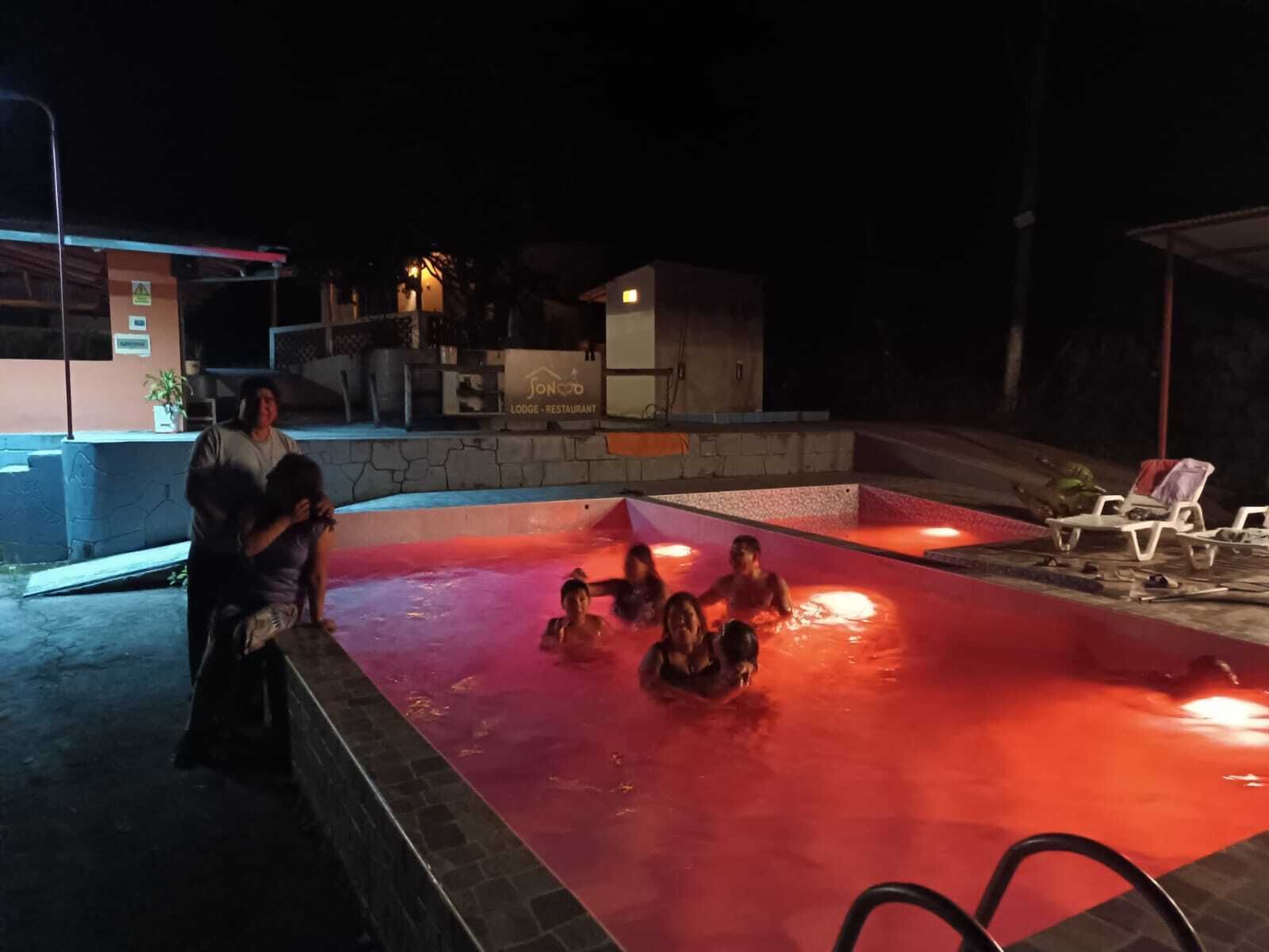 A swimming pool area at night with a red-lit water feature, lounge chairs, and a building labeled 'SONO LODGE RESTAURANT' in the background.