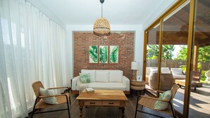 A living room with a white sofa, two wicker chairs, a wooden coffee table, and a brick wall with leaf paintings.