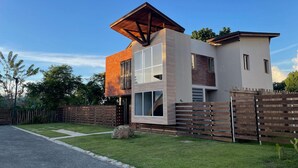 A modern two-story house with a flat roof, large windows, and a wooden fence.