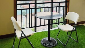 Una habitación con un ventanal grande, dos sillas blancas, una mesa redonda y alfombra verde sintética.