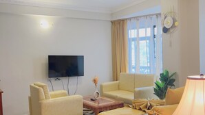 Habitación de hotel con cama, dos sillones, sofá, televisor, ventana con cortinas y un reloj de pared.