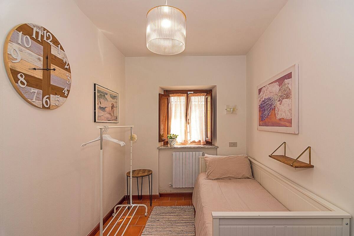 A small, cozy bedroom with a bed, a radiator, a window with curtains, a hanging light fixture, and a wall clock.