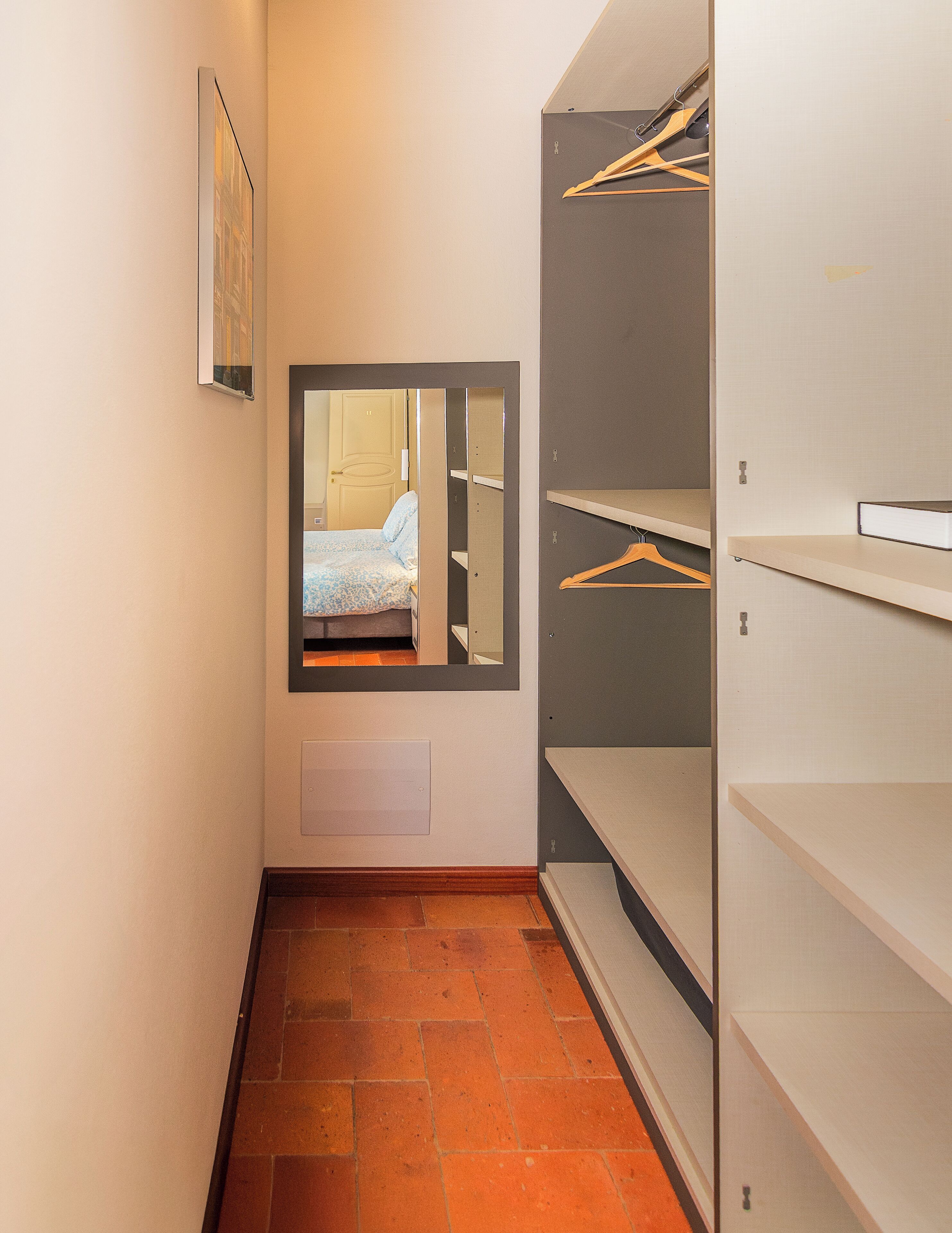 Een smalle gang met een bed op de achtergrond, een spiegel en een garderobe met kledinghangers.