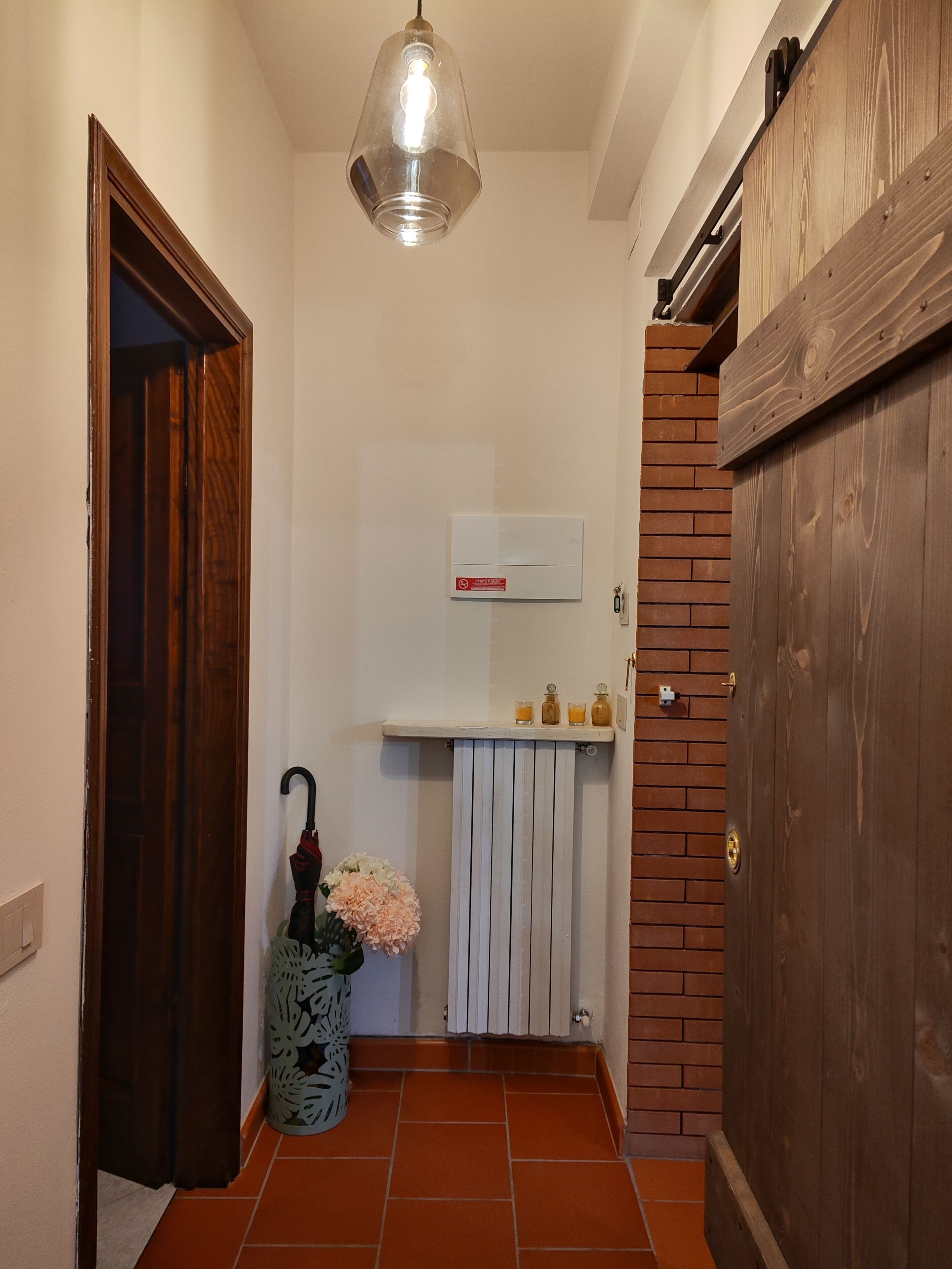 Un couloir avec une porte en bois, un radiateur, une étagère avec des bouteilles et un luminaire suspendu.