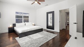 A modern bedroom with a large bed, a ceiling fan, a window with blinds, a mirror, and a door leading to another room.