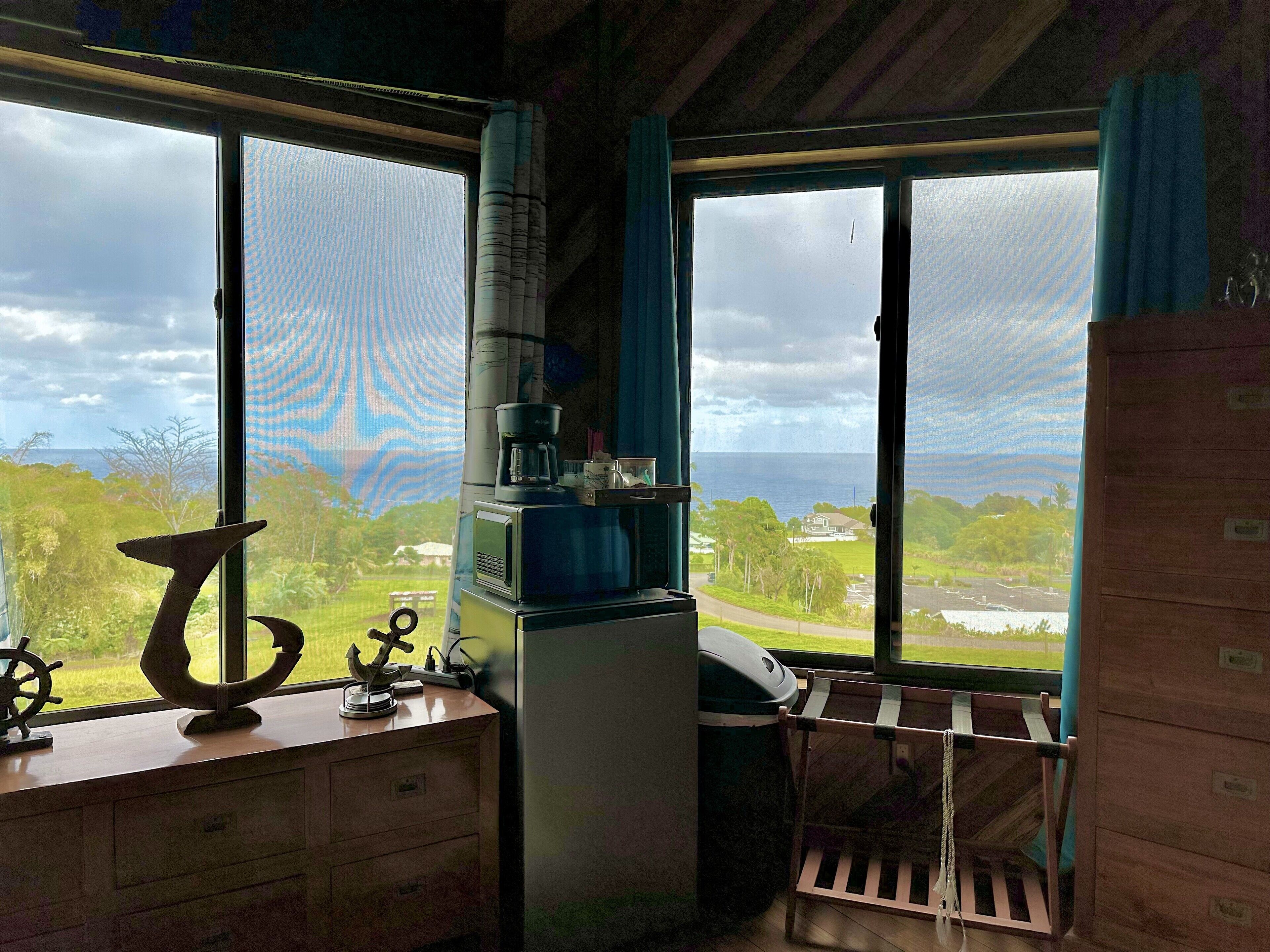 A room with a view of the ocean and a window with curtains. There is a desk with a coffee maker and a chair.