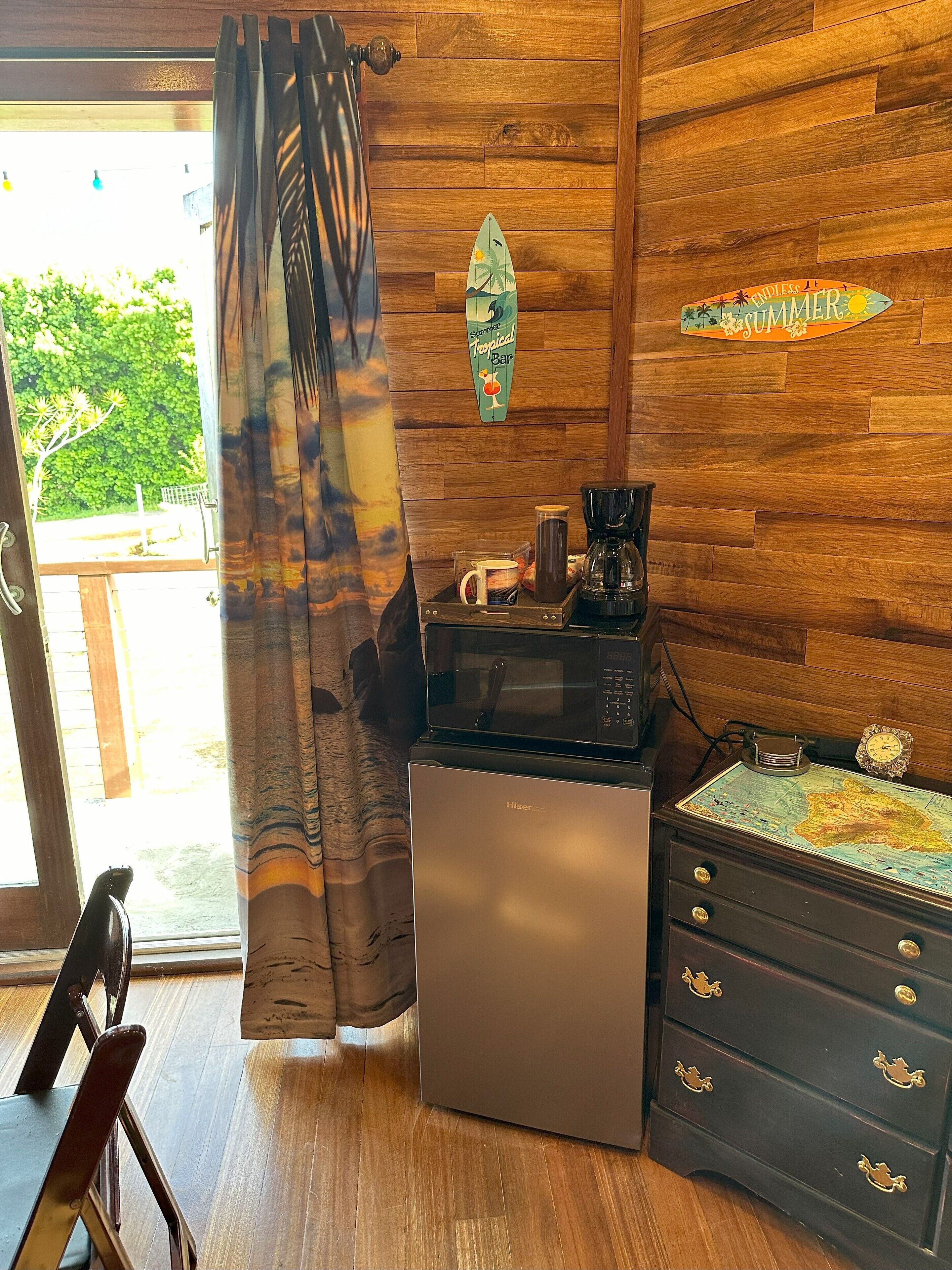 A room with wooden walls, a wooden floor, a microwave, a coffee maker, a small refrigerator, a cabinet with drawers, a map, and a sign that says "Endless Summer Bar".