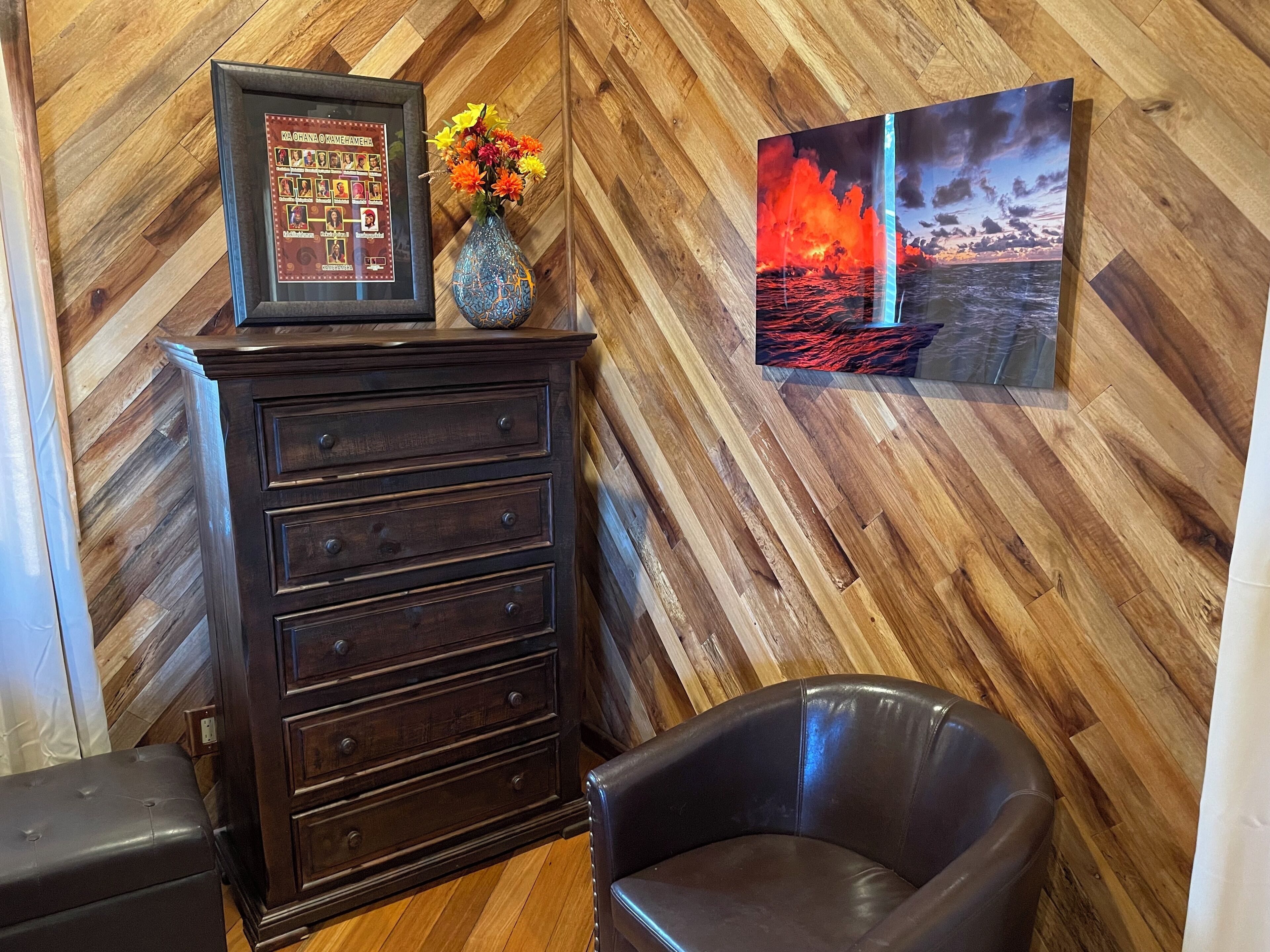 Un pavimento in legno a spina di pesce, un comò scuro con cassetti, un vaso con fiori e un quadro raffigurante un flusso di lava.
