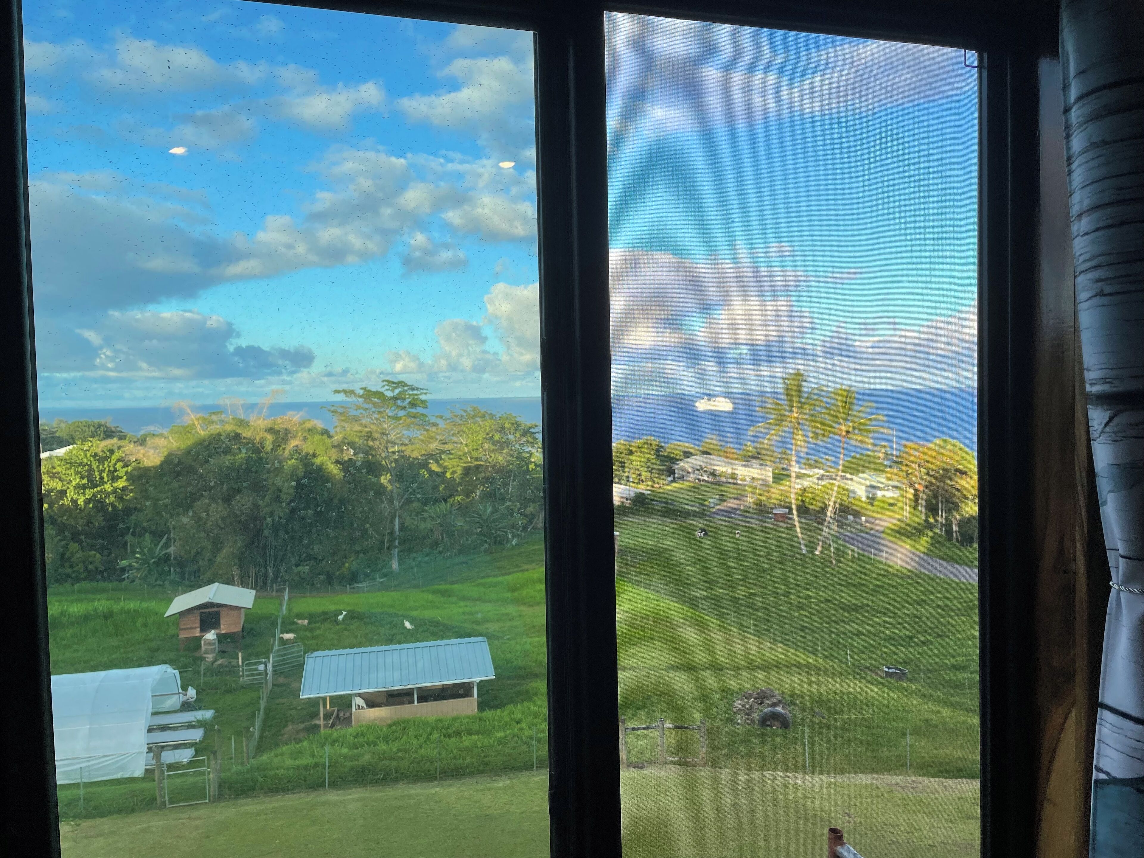 A view through a window with a rural landscape, including a grassy field, a small building, and a road with a vehicle.