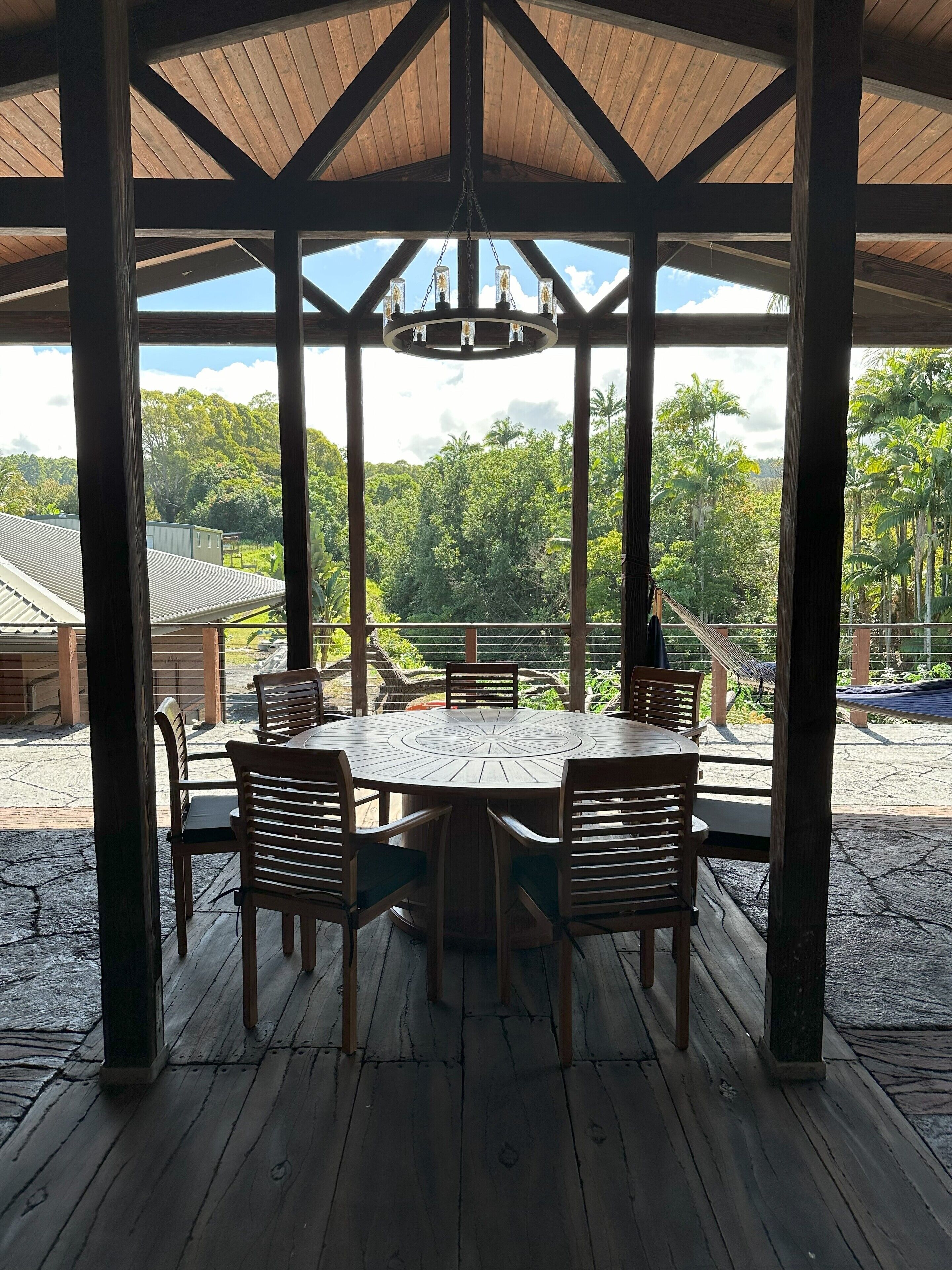 A covered outdoor dining area with a round wooden table and chairs, a hanging light fixture, and a view of lush greenery and buildings in the background.