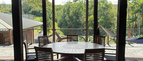 A covered outdoor dining area with a round wooden table and chairs, a hanging light fixture, and a view of lush greenery and buildings in the background.
