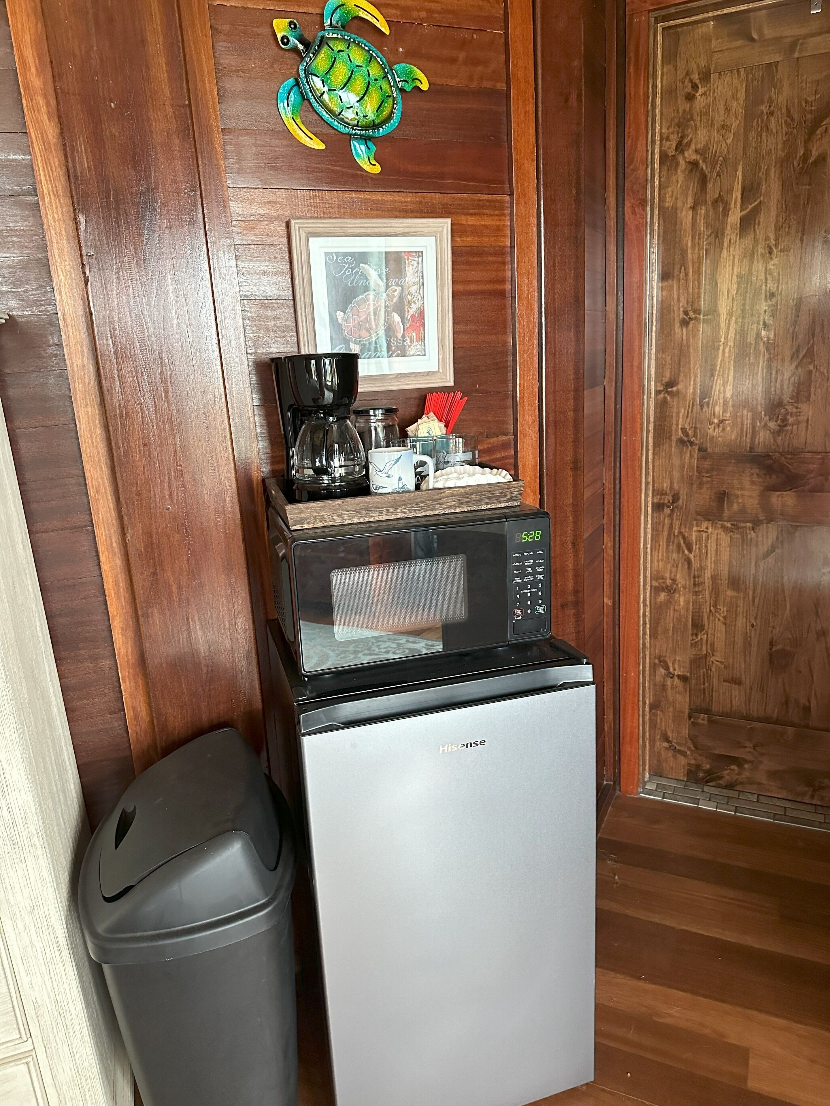 A compact kitchen area with a microwave, coffee maker, and a trash bin. A framed picture hangs on the wall above the microwave.