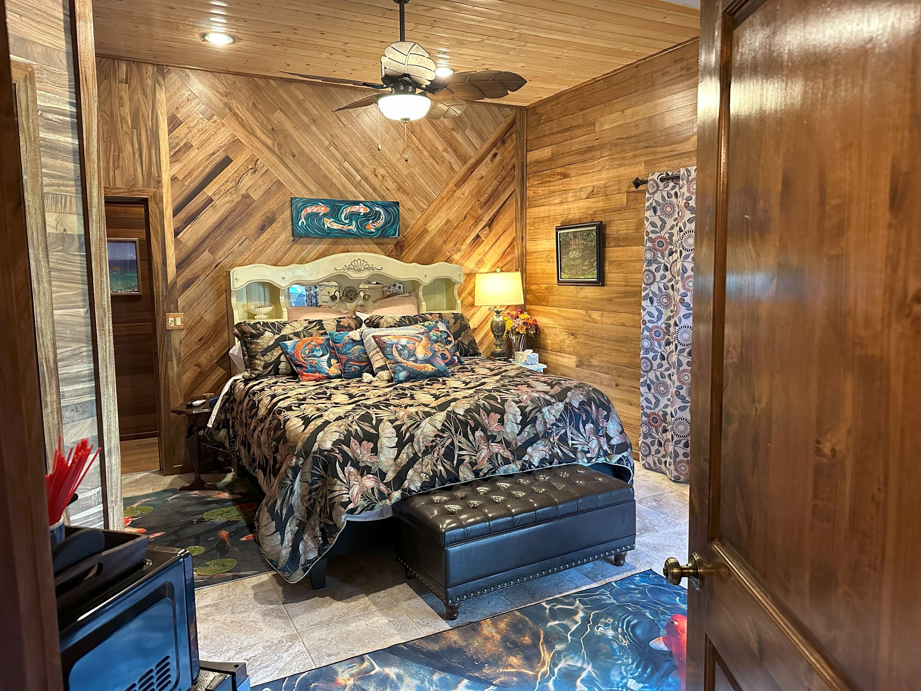 A bedroom with a wooden ceiling, a bed with floral bedding, a nightstand, a lamp, and a decorative wall piece.
