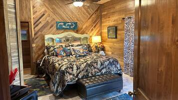A bedroom with a wooden ceiling, a bed with floral bedding, a nightstand, a lamp, and a decorative wall piece.