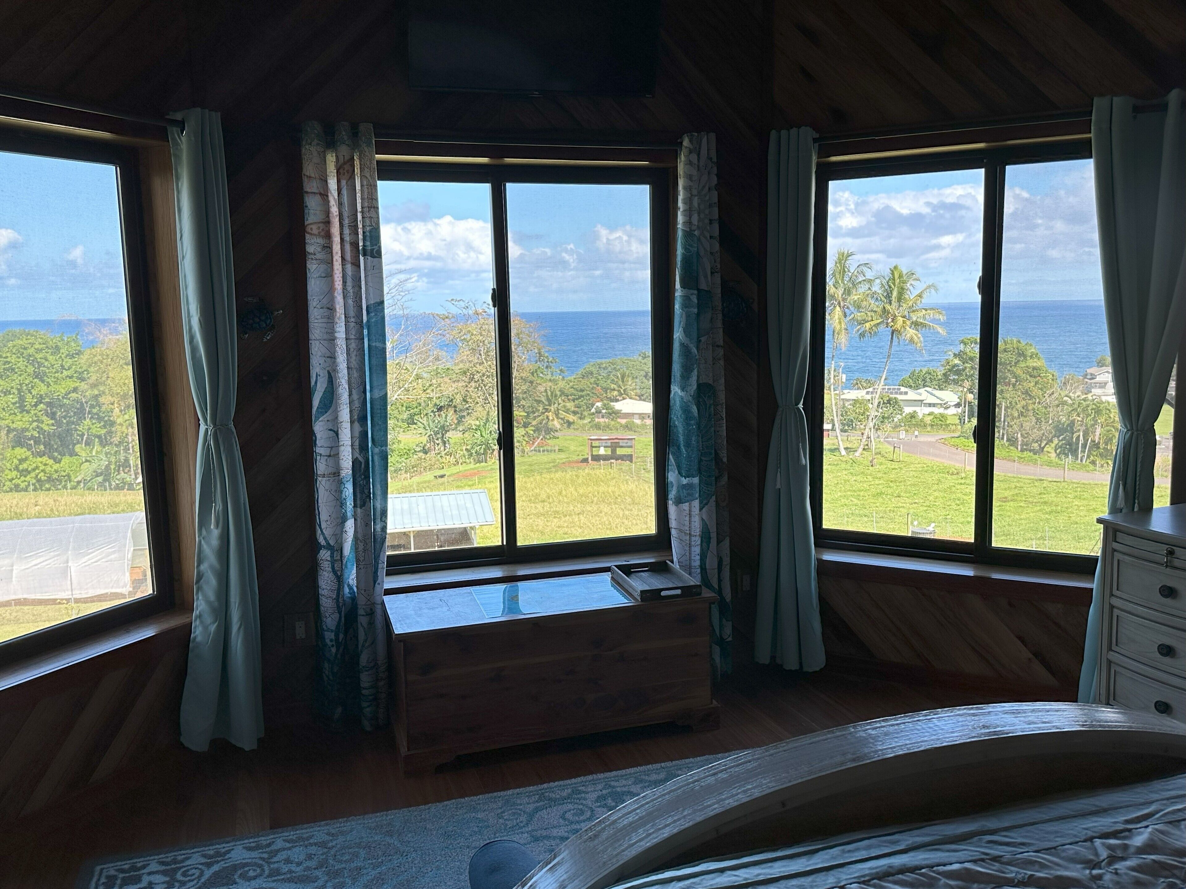 A room with a wooden floor, a bed, a wooden bench, and large windows offering a view of the ocean and greenery.