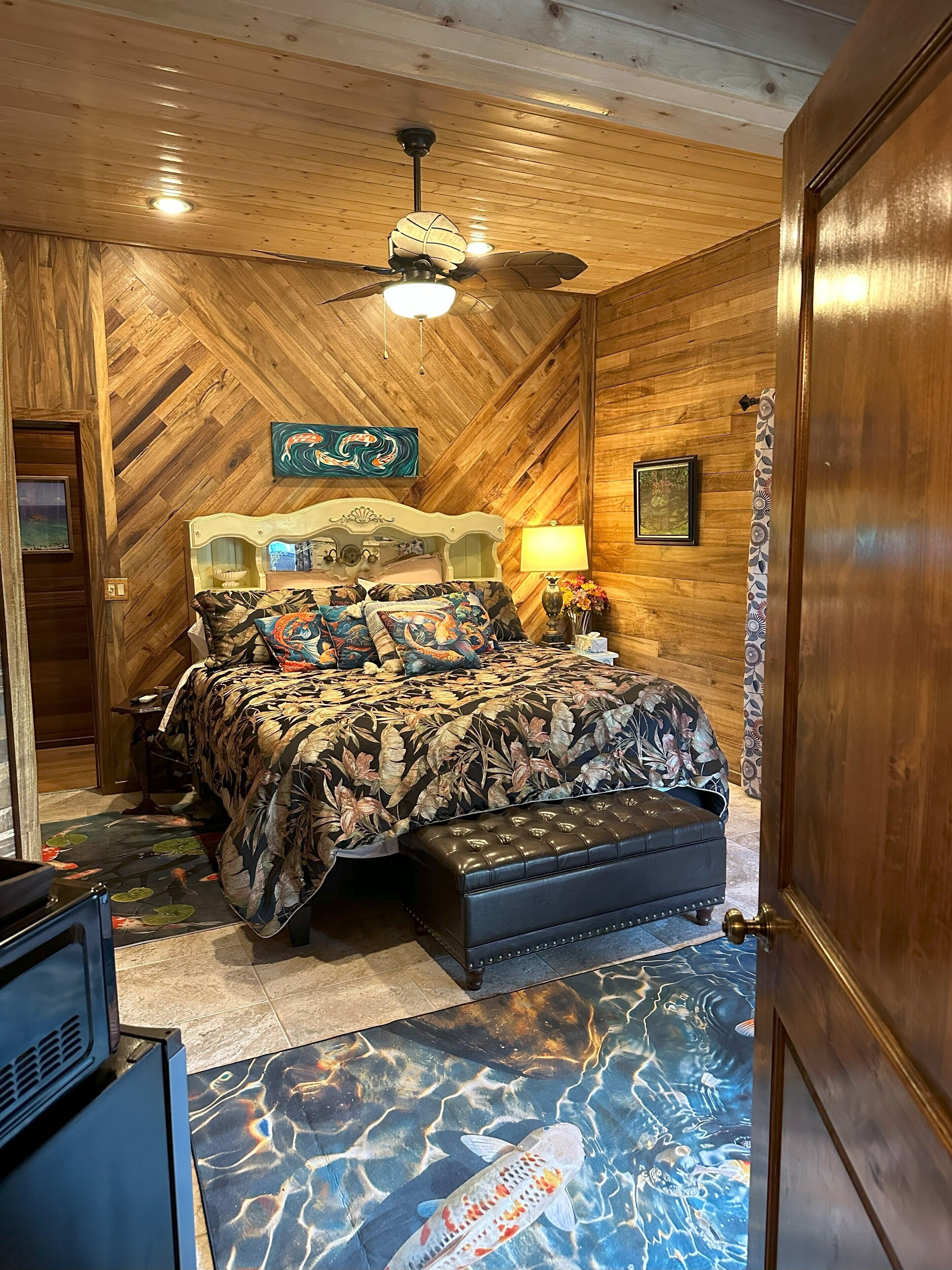 A bedroom with a wooden ceiling, a bed with a floral bedspread, a ceiling fan, and a wall-mounted lamp.