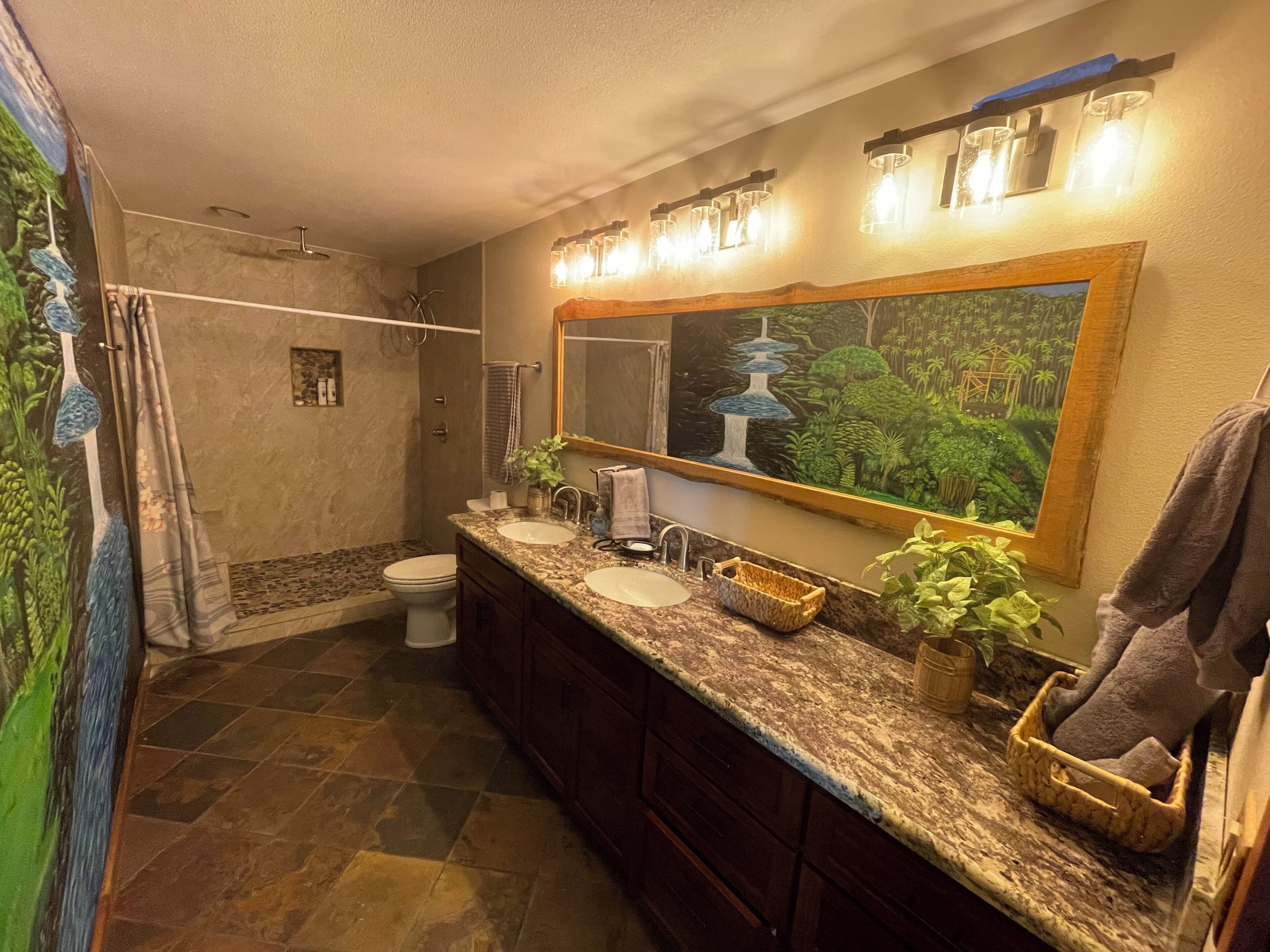 A bathroom with a shower, two sinks, a large mirror, and a painting of a waterfall.