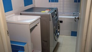Kompakti pyykinhuoltotila, jossa on pyykinpesukone ja -kuivausrumpu, pieni hylly ja laatoitettu lattia.