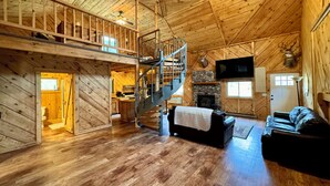 A wooden cabin interior with a loft bedroom, a stone fireplace, a staircase, and a mounted television.