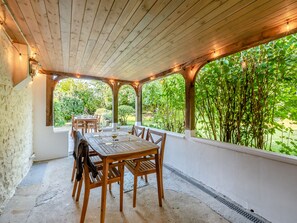 A covered patio with a wooden table and chairs, string lights, and a view of greenery. - Charming four-bed holiday home in Yarcombe sleeps eight. (Yarcombe)
