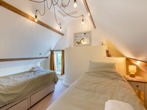 A bedroom with two beds, a window, a bedside table with a lamp, and a ceiling with exposed beams and light fixtures. - Charming four-bed holiday home in Yarcombe sleeps eight. (Yarcombe)