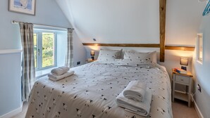 A bedroom with a bed, a nightstand, a lamp, a window with curtains, and a framed picture on the wall. - Charming four-bed holiday home in Yarcombe sleeps eight. (Yarcombe)