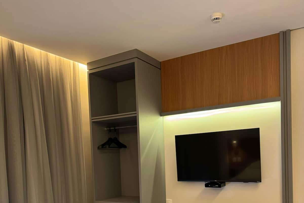 Una habitación de hotel moderna con escritorio integrado, televisor de pantalla plana montado en la pared, armario con perchero y refrigerador compacto.
